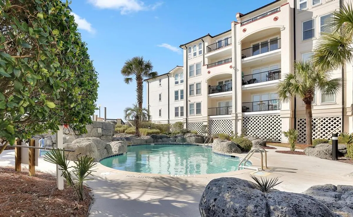 A close-up exterior view of the sparkling swimming pool surrounded by lush palm trees, landscape rocks, and the apartment building. Sunbathers and guests are gathered around the pool, enjoying the tranquil and refreshing environment. The scene highlights the inviting pool deck and the sense of community at the heart of the amenities.