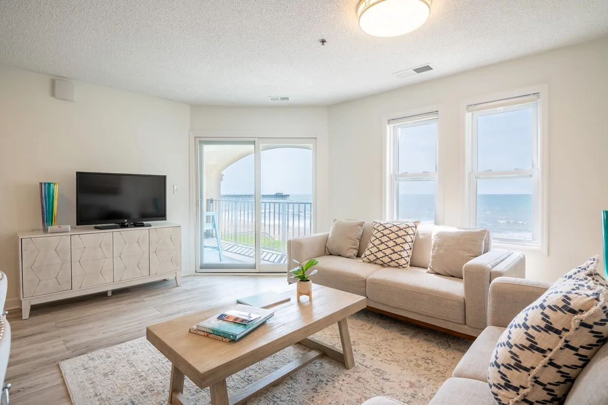 A cozy living room featuring a plush sofa, coffee table, and a flat-screen television. Large sliding doors open directly onto the balcony, framing breathtaking ocean views and letting in abundant natural light.