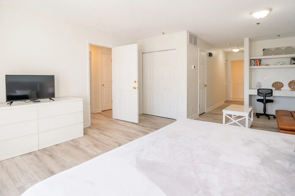 A serene primary suite with a comfortable bed, a desk area suitable for remote work or reading, a wall-mounted television, and open doors leading to natural light. The neutral color palette and functional layout create a peaceful retreat for adults.