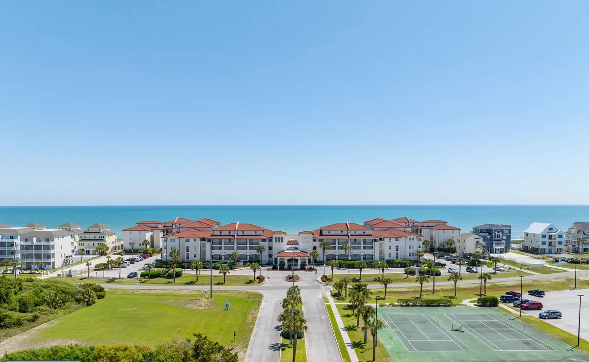 A vibrant exterior shot featuring the oceanfront property with immediate beach access, inviting hotel architecture, tennis courts, and lush landscaped gardens. The beach is visible with families engaged in activities like swimming, playing beach volleyball, and relaxing by the pool.