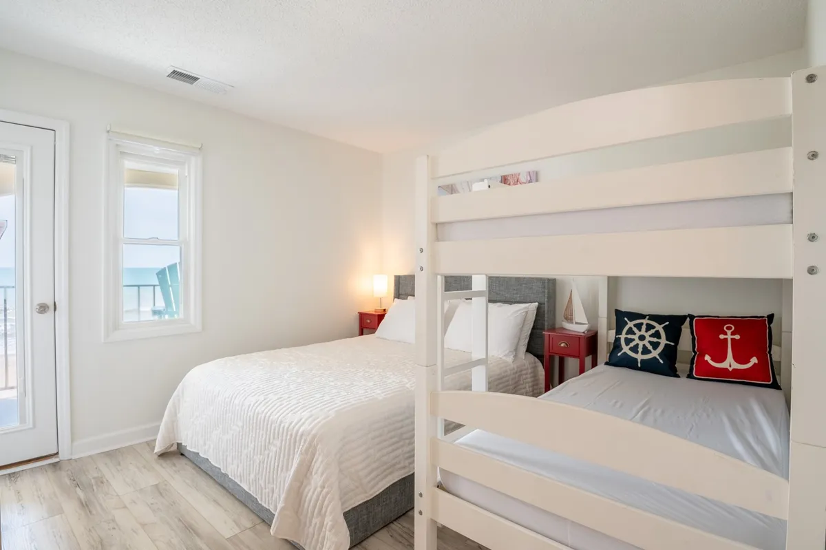 A spacious bedroom featuring a queen bed, full-over-full bunk beds, nautical-themed pillows, and large windows that offer ocean views. Nightstands with lamps flank the beds, creating a cozy, family-friendly atmosphere ideal for group stays or children's sleepovers.