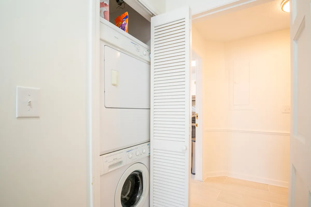 A tidy, well-lit laundry closet equipped with a stacked washer and dryer, white cabinetry, and light-colored walls. The setup is practical and organized, demonstrating the convenience of on-site amenities for longer stays and family travel.