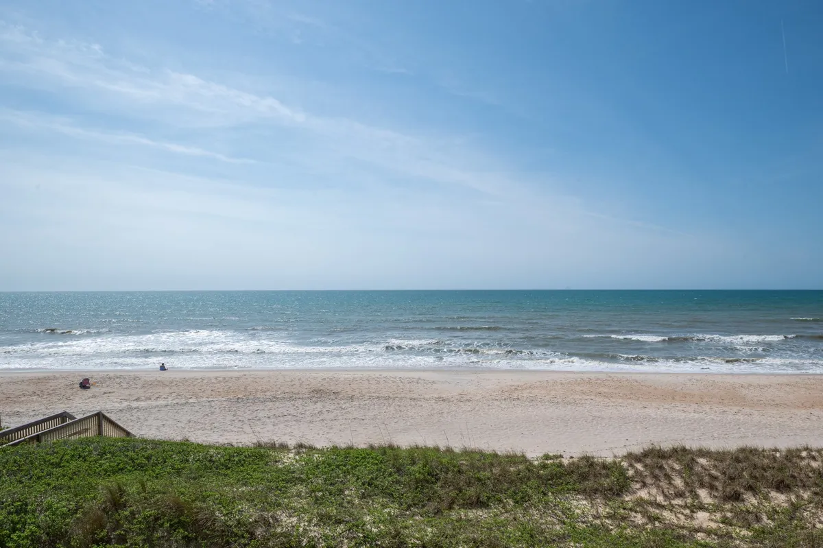 A scenic view of the beach with soft dune grass in the foreground, gentle waves, and people sunbathing, swimming, or strolling along the shore. The sky is clear, evoking a peaceful and inviting seaside atmosphere.