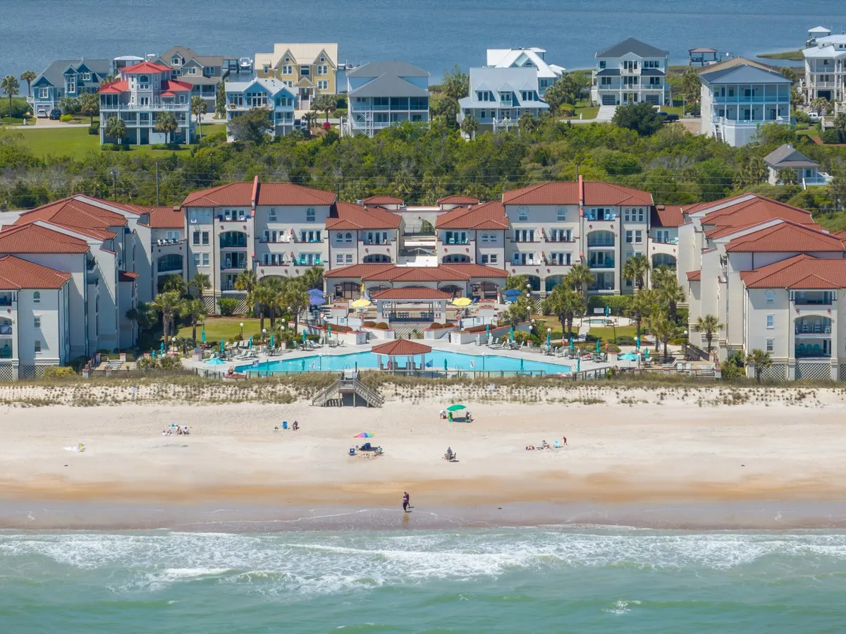A spacious exterior view of the resort complex, featuring the central swimming pool, surrounding palm trees, beachfront, ocean, and nearby houses. The pool takes center stage, with lounge areas and sunbathers contributing to a lively, vacation-ready atmosphere. The ocean and beachfront are visible beyond the pool, emphasizing easy private beach access.